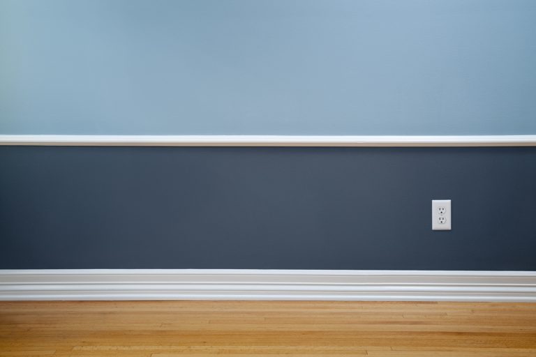 A light and dark blue colored wall with a white colored baseboard with wooden laminated flooring, What is the Standard Size of a Baseboard? [And Why]