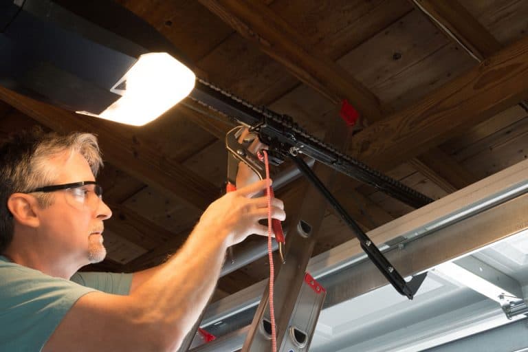 Closeup of a professional automatic garage door opener repair service technician man working on a ladder at a home residential location making adjustments and fixing it while installing it, How To Close A Garage Door Manually
