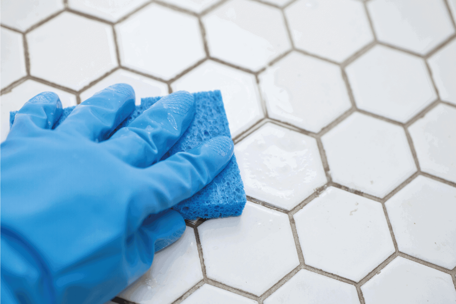 a person wearing blue gloves holds a sponge cleaning a white tile floor. Should You Seal Floor Grout [Breakdown By Room And Type Of Grout]