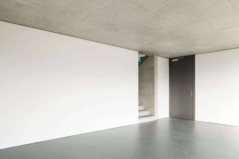 Interior of an empty living room with concrete flooring, white walls, and gray ceiling, How Thick Does A Concrete Floor Need To Be?