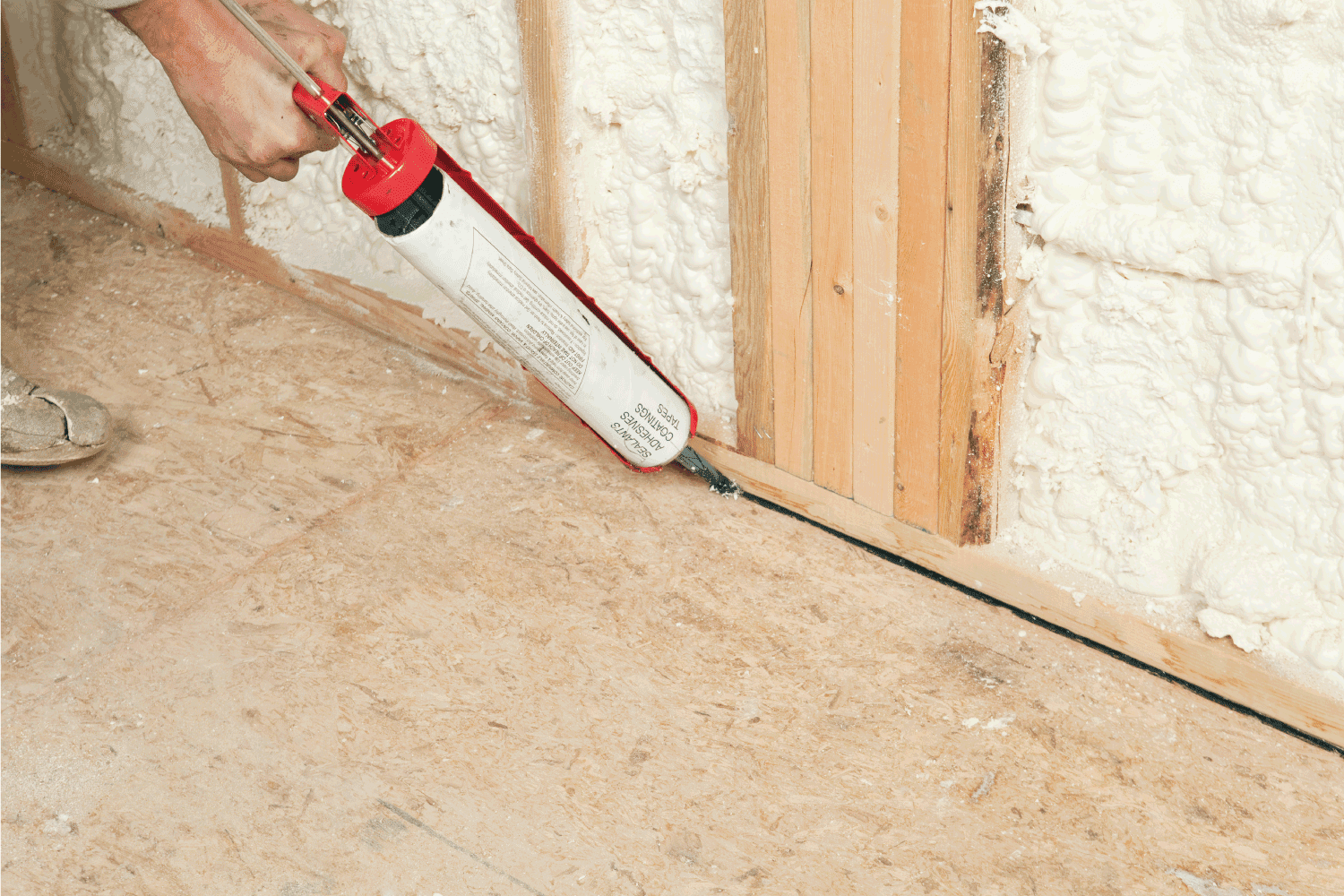 A construction worker is applying butyl sealant caulk to gaps between wall plate and OSB subfloor at a house