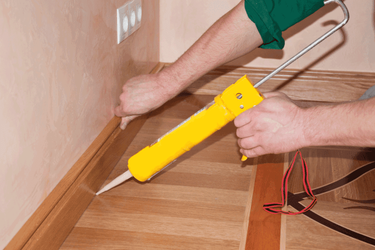 Oak-Wood-floor-installation.-Carpenter-on-work-putting-wood-parquet-skirting-board-with-glue.-How-To-Fill-A-Gap-Between-Wood-Floor-And-Wall