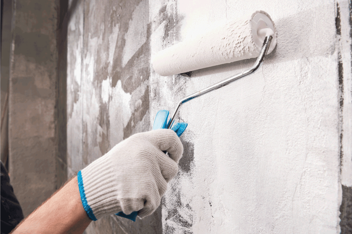 man wearing work gloves and using roller to paint a wall white. How To Paint The Foundation Of Your House