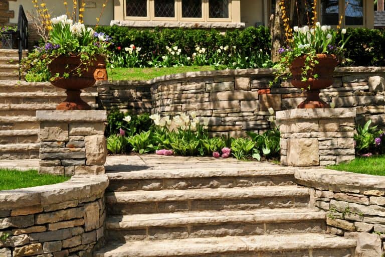 A gorgeous rock landscape in the front of the house with rock staircase and retaining walls decorated with flowers, How Close Can A Retaining Wall Be To A House Foundation?