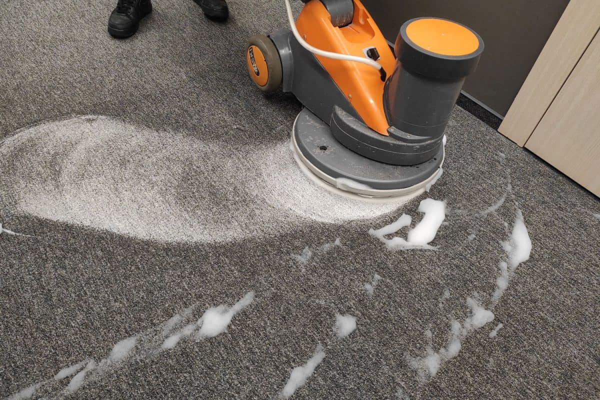 A worker cleaning the gray carpet using a disk cleaner