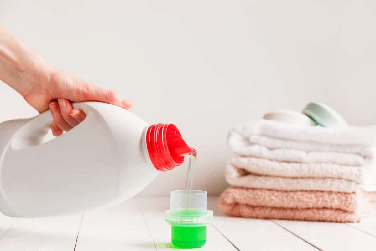 Hands pouring liquid laundry detergent into cap on white rustic table with towels on background in bathroom