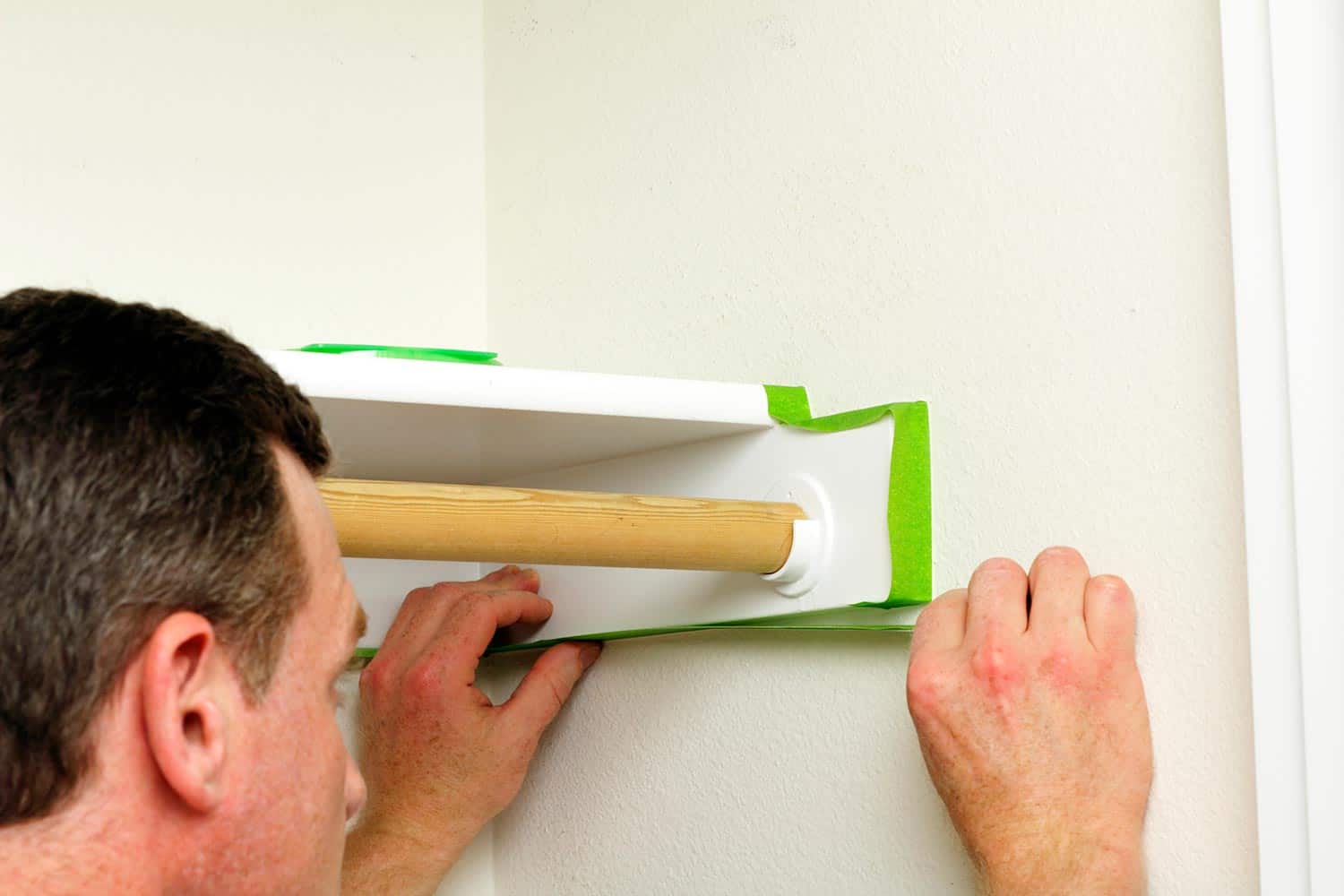 A man is preparing to paint the closet walls by putting on green painter's tape to the edges around a shelf