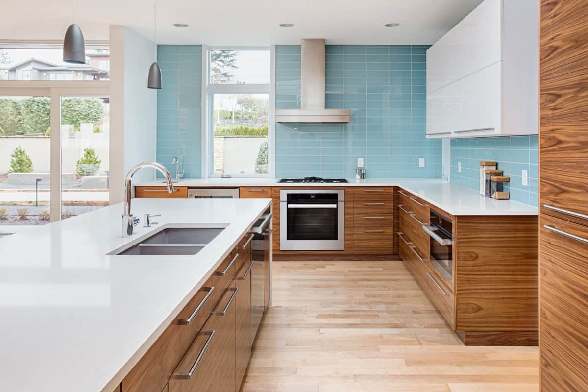 A spacious and brightly lit kitchen with white countertops, blue backsplash and wooden cabinets and white cupboards