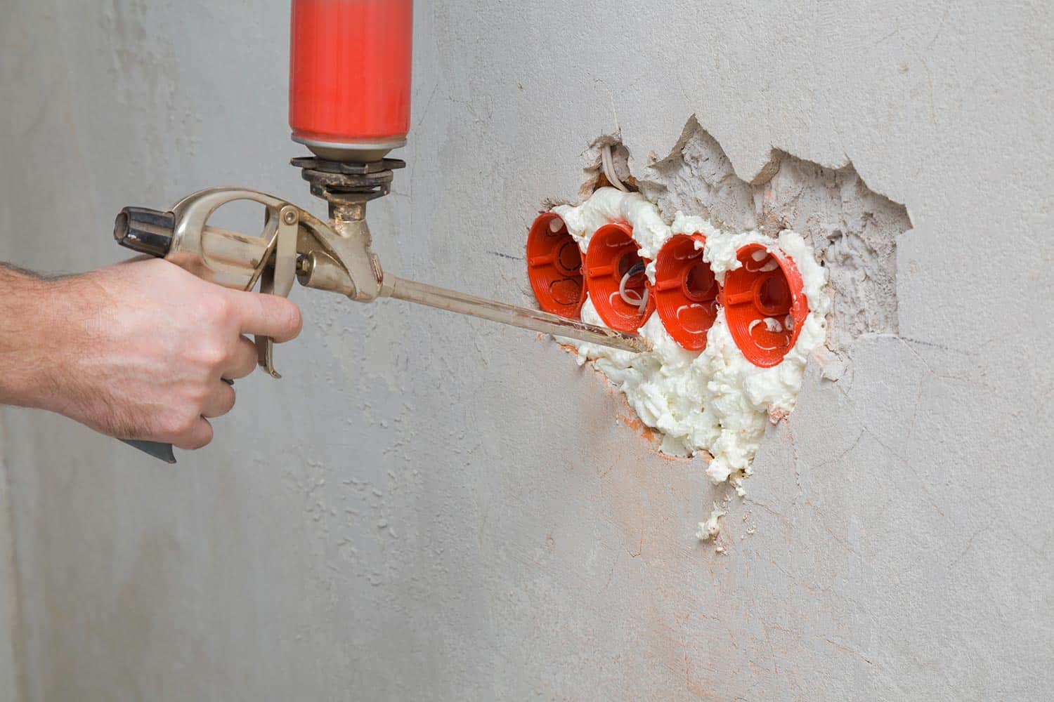 Young adult man hand using spray gun and filling gap with construction foam between new plastic red electric outlet and concrete wall