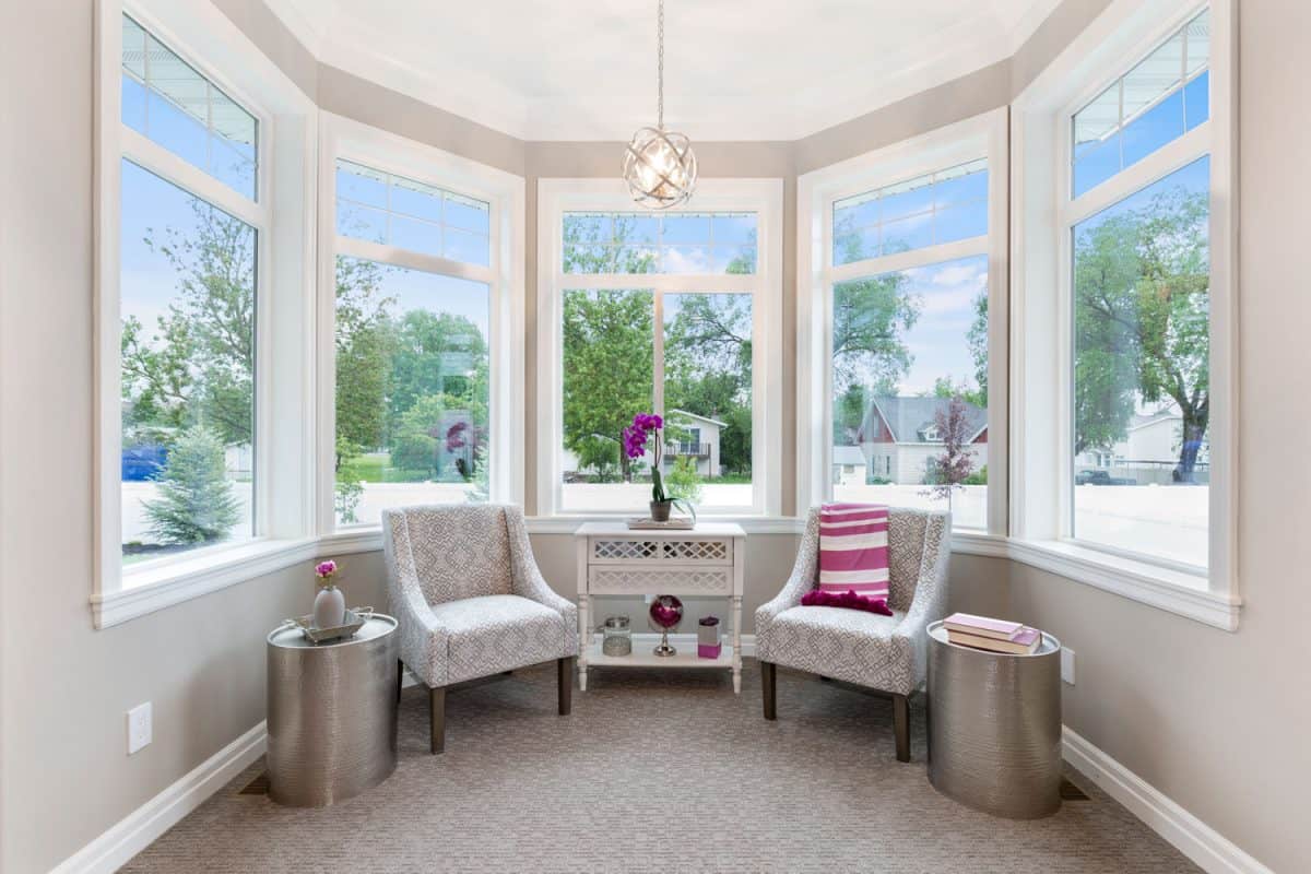 A bay window with frameless windows with carpeted flooring, gray armchairs and a lavender on the end table