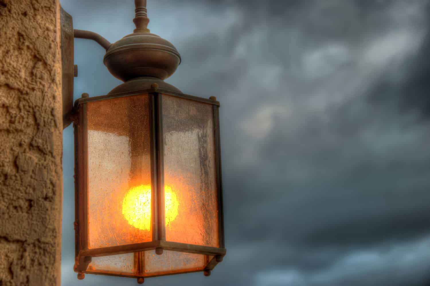 Solitary outdoor exterior wall mounted electrical lantern shining brightly in its glory of red, yellow, orange light against the background of heavily clouded, storm gathering sky.