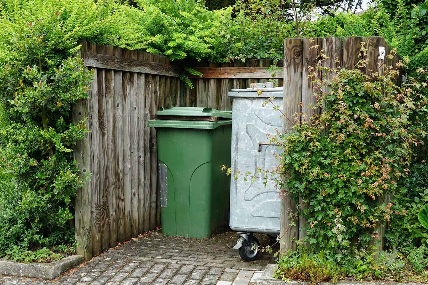 Metal container for house waste and plastic container for organic waste in a wooden niche surrounded by lush vegetation