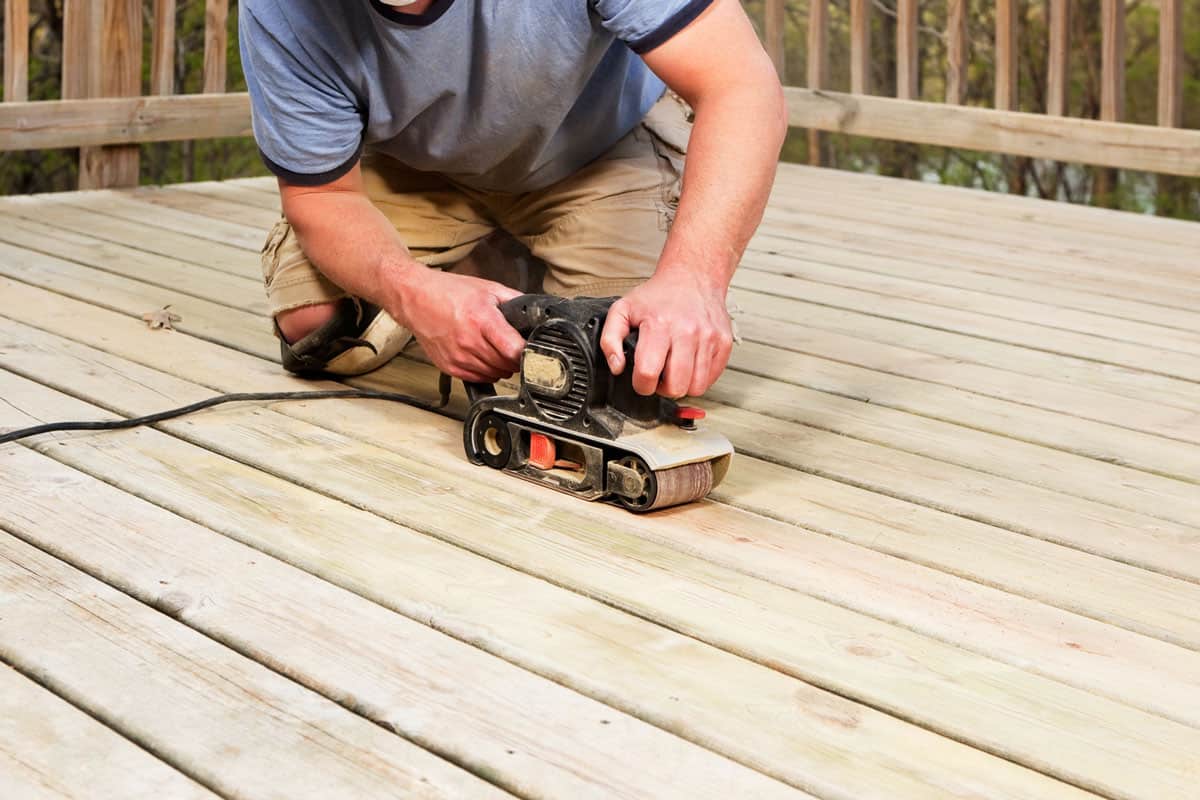 A worker is using a belt sander to sand and recondition weathered deck boards in preparation for staining
