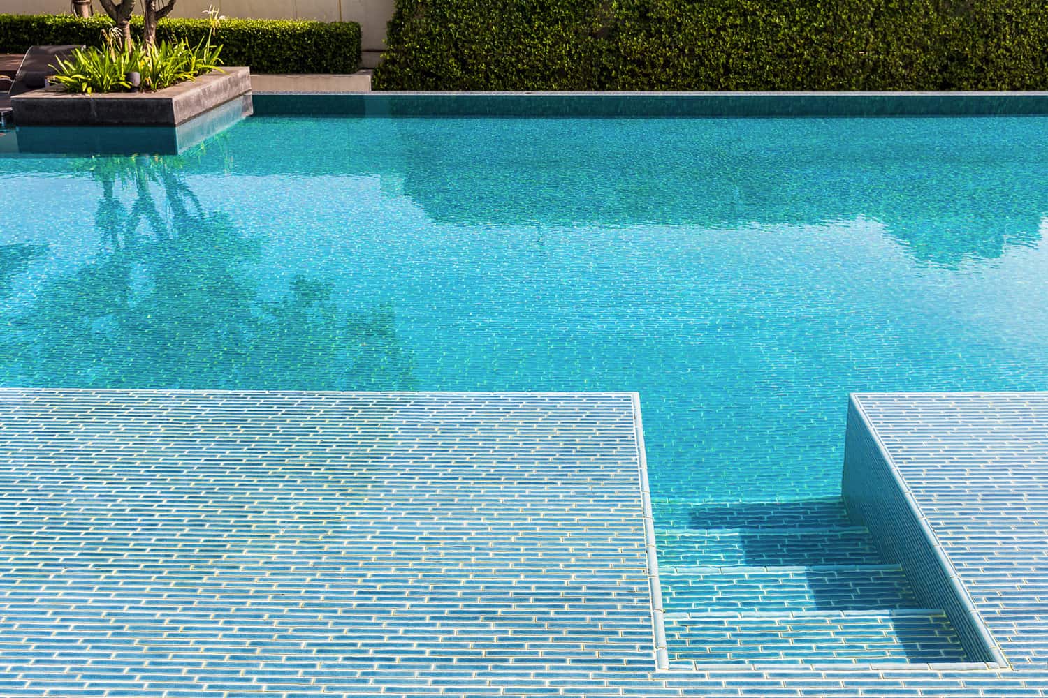 Swimming pool made by mosaic tiles with small stairs inside the clear blue water which not moving. Water surface reflects shadow of sky, tree and bush in far background.