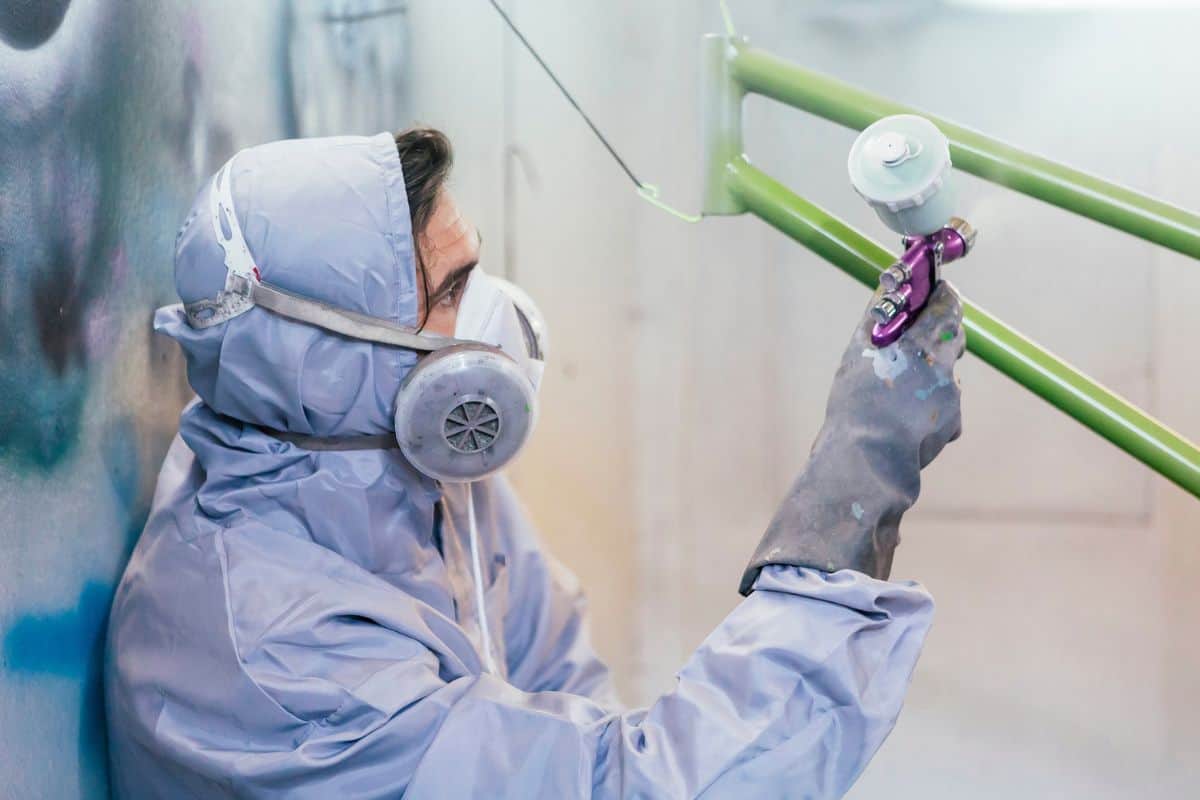 Side view of man in uniform and respirator painting bikes frame with green color