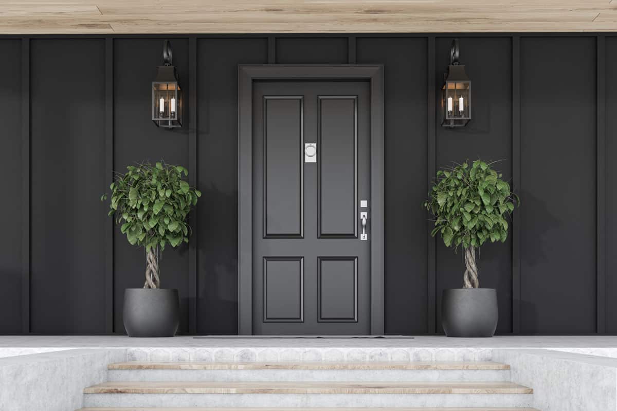 Stylish black front door of modern house with black walls, door mat, trees in pots, stairs and lamps