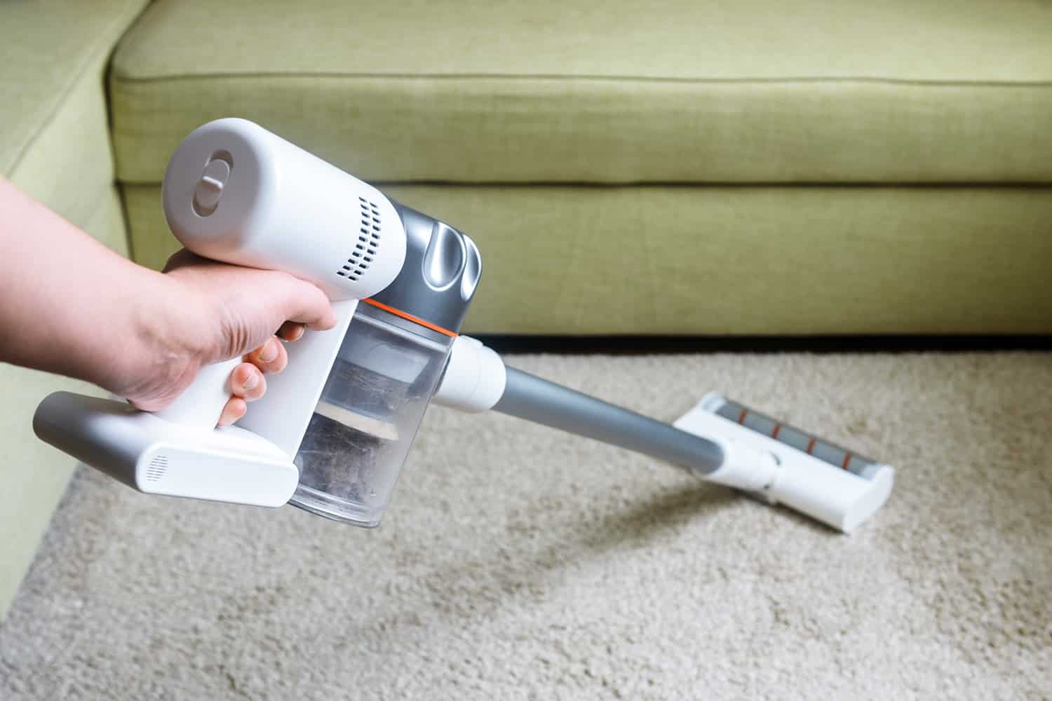 Wireless vacuum cleaner used on carpet in room. Housework with new handy upright stick hoover. Person holds vacuum cleaner, modern white broom by sofa and floor. Home cleaning and care concept.