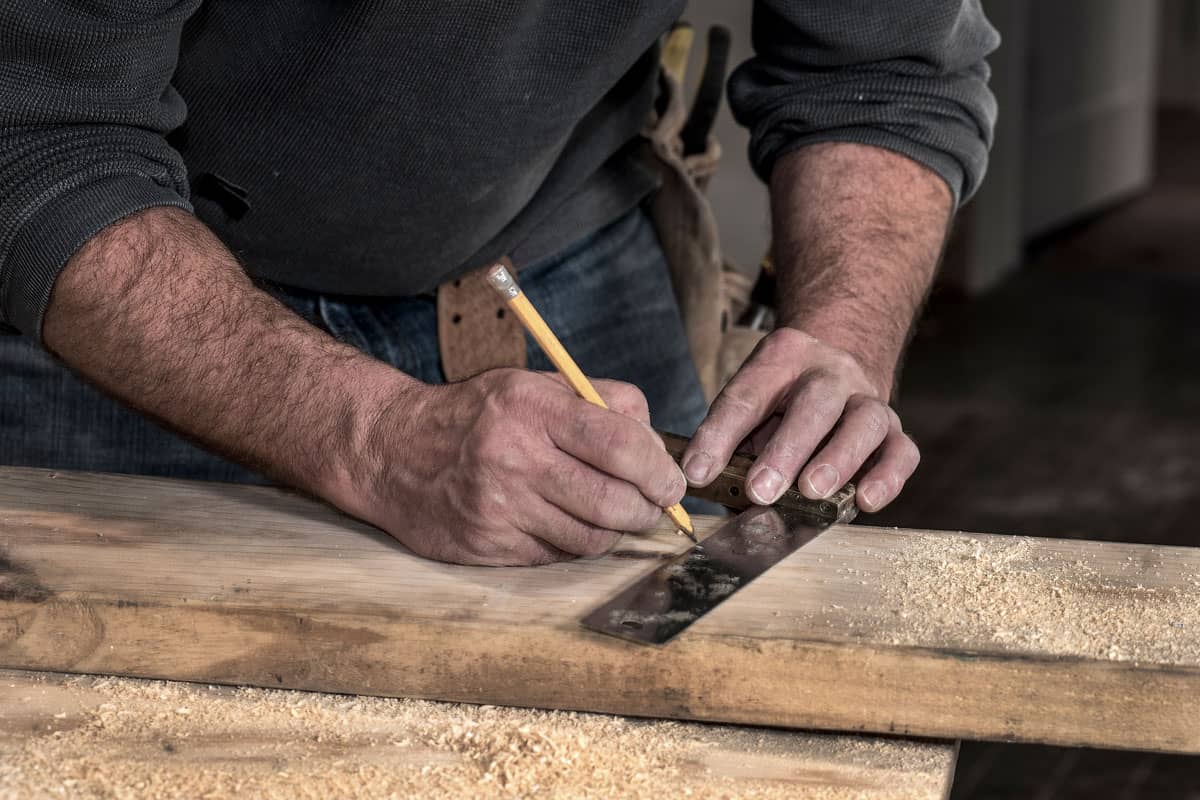 carpenter, tool belt, and rough hands using pencil and old wooden handle square to mark line on a wood board to cut, during