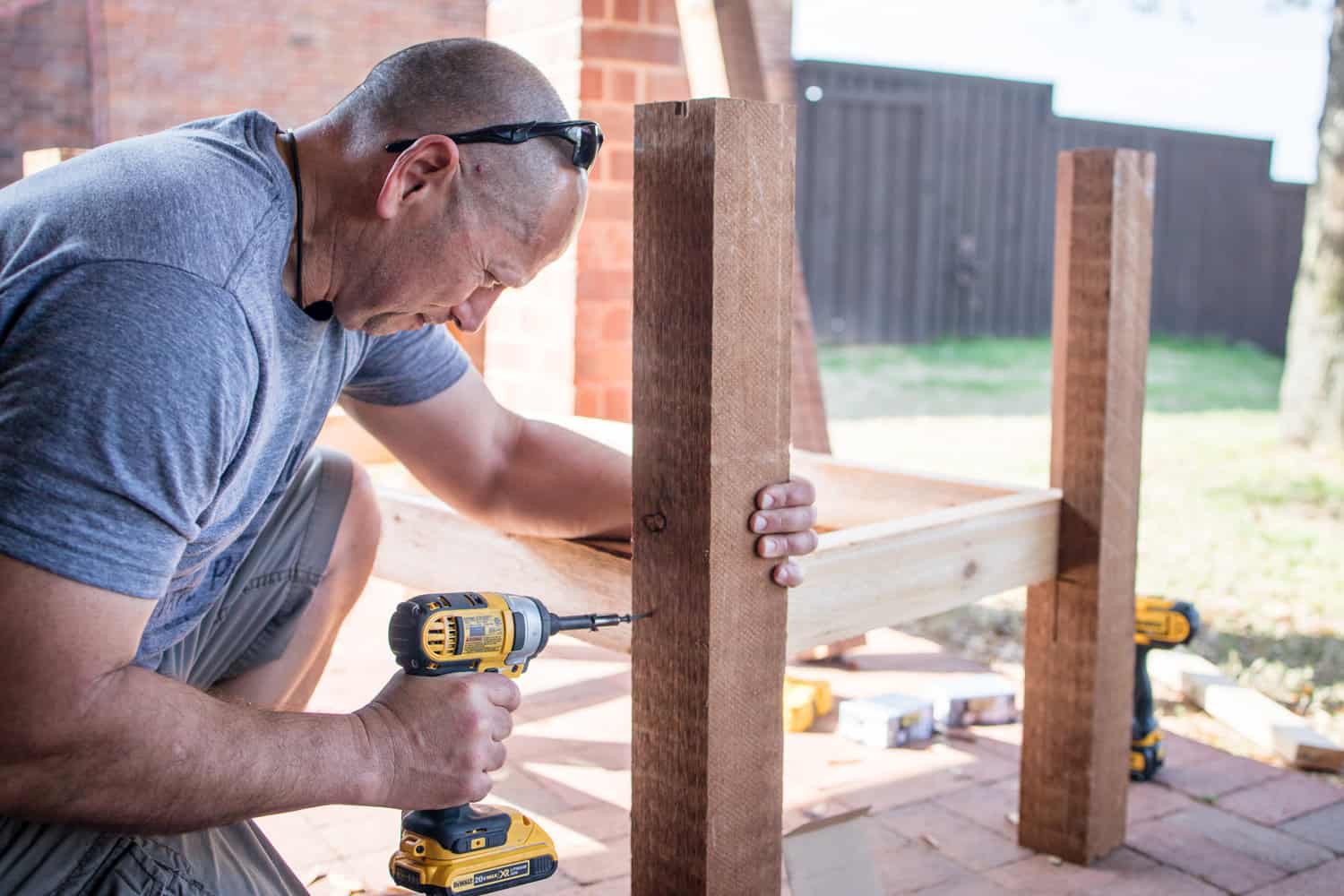 Side view of a DIY man as he uses a cordless electric power screwdriver to place screws into a wooden post while building a backyard container garden planter.