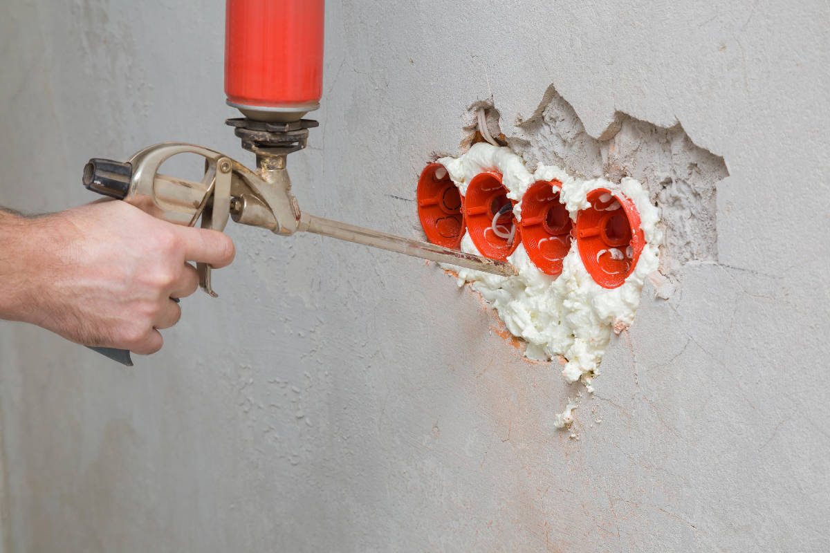 Young man hand using spray gun and filling gap with construction foam between new plastic red electric outlet and concrete wall