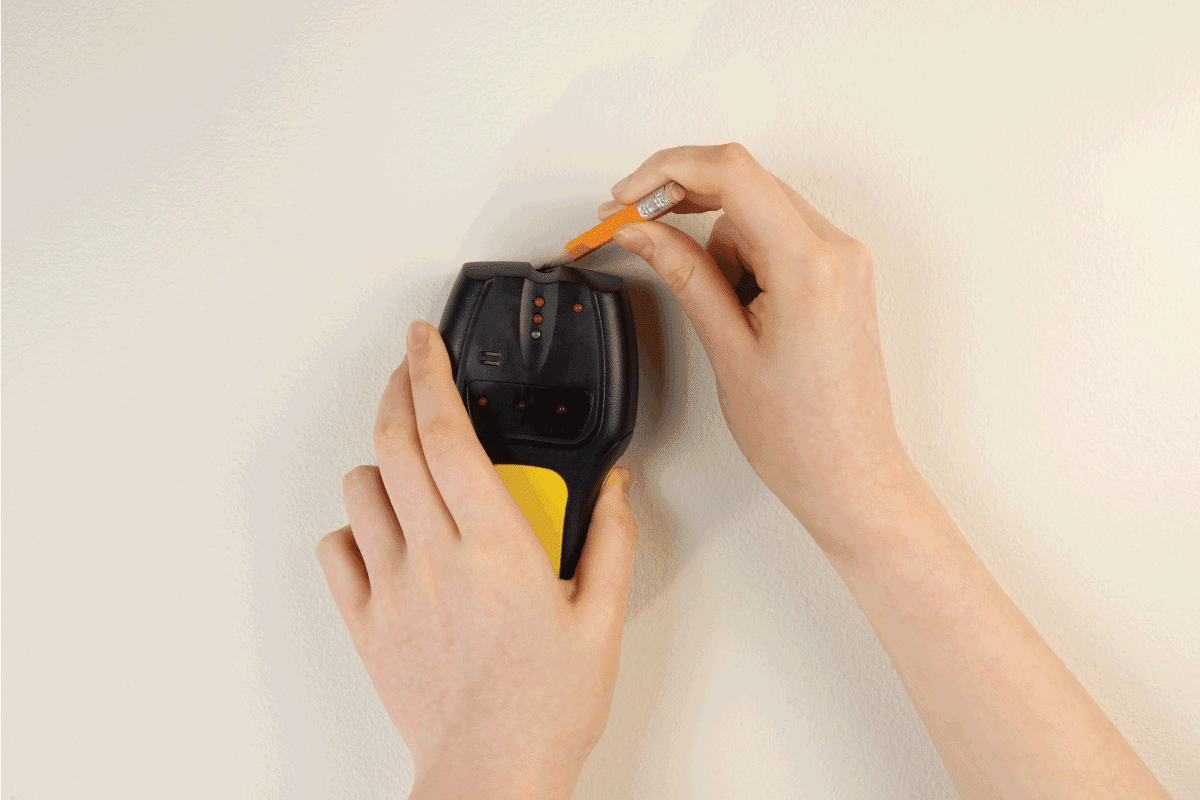 Hands holding using stud finder and pencil against interior home white wall, How To Use A Tavool Stud Finder [Step By Step Guide]