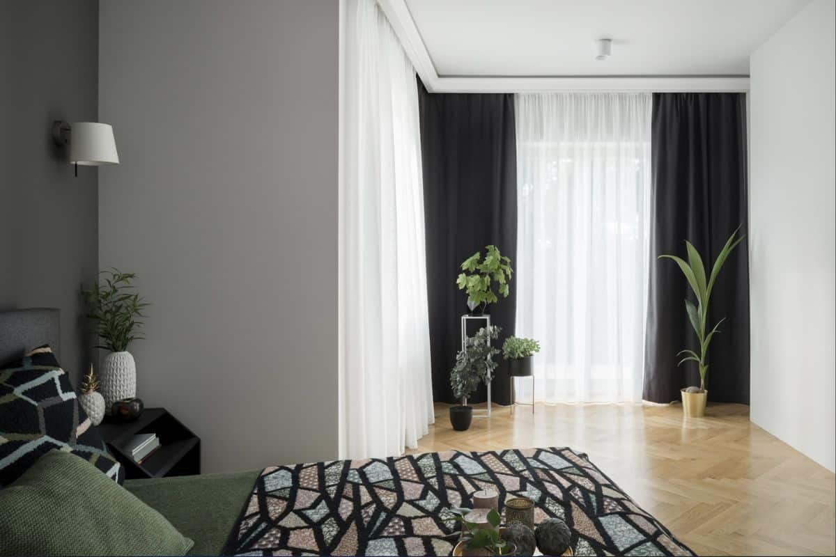 Comfortable and stylish bedroom with big bed, black bedside table with decorations, nice wooden floor and big window walls behind long curtains.