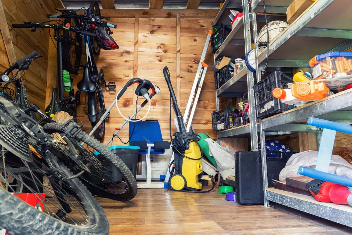 Suburban home wooden storage utility unit shed with miscellaneous stuff on shelves, bikes, exercise machine, ladder, garden tools and equipment. Messy and chaos at house yard barn.