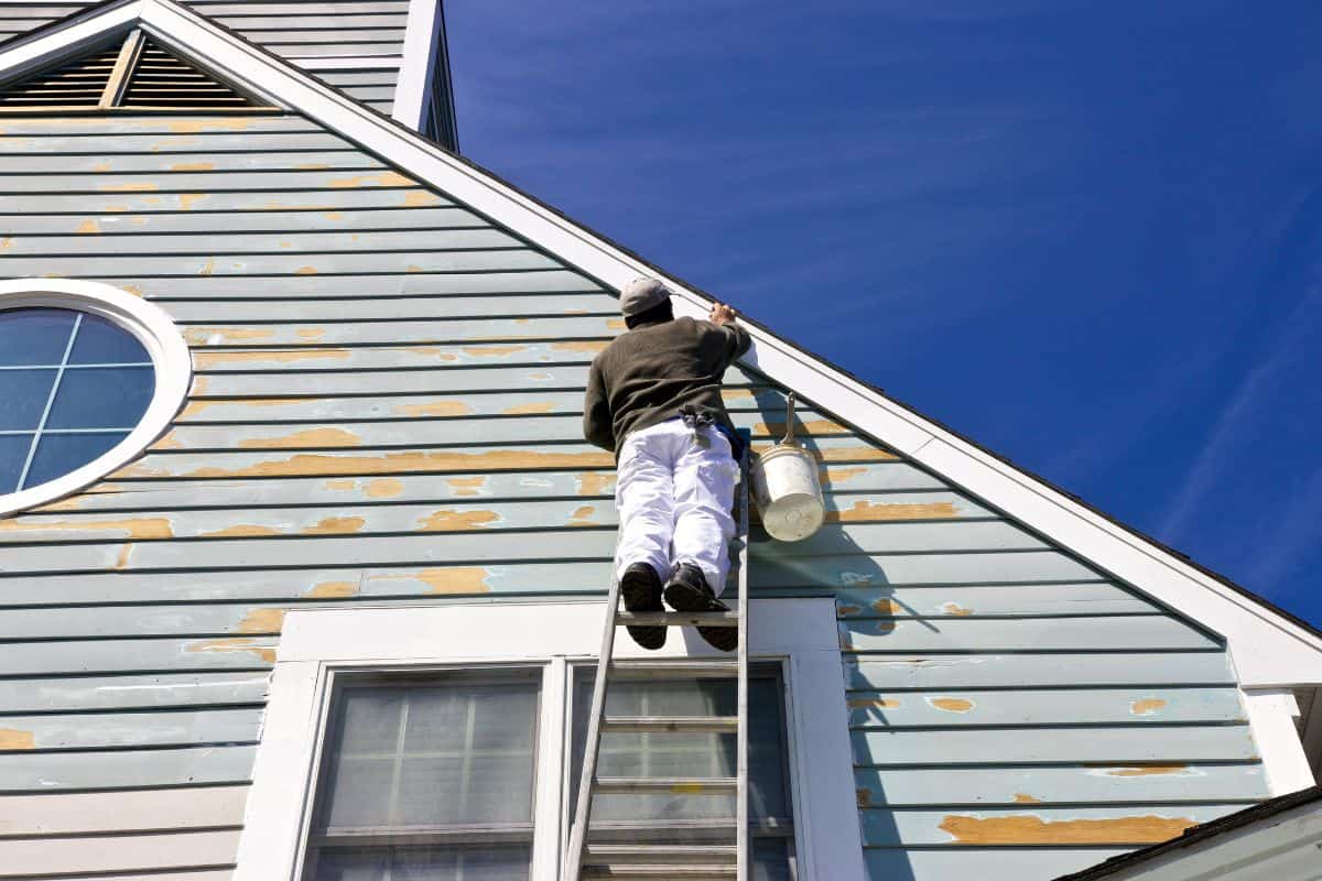 A contractor or painter on a ladder doing exterior paint work, sanding, trim work and repairs on a house, condo or building with wood siding. Blue sky background.