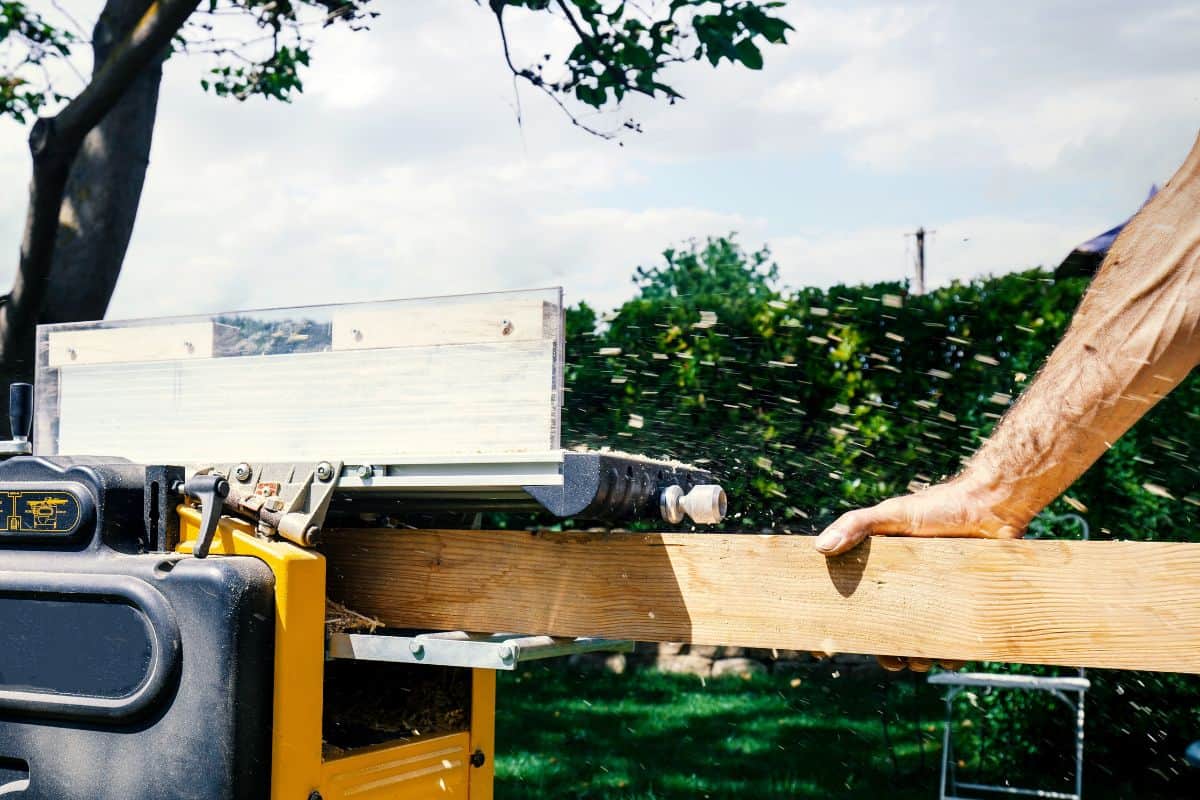 Joiner, Man's hand scouring a wooden board on a thickness machine in garden. Carpenter working with electric planer on wooden beam, plank