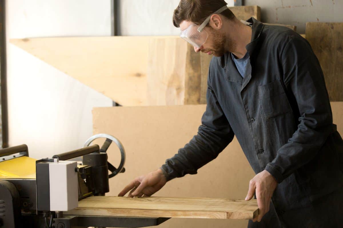 Professional woodworker in protective goggles making wooden parts for custom furniture on machine tool. Workman working at thickness planer machine in carpentry.