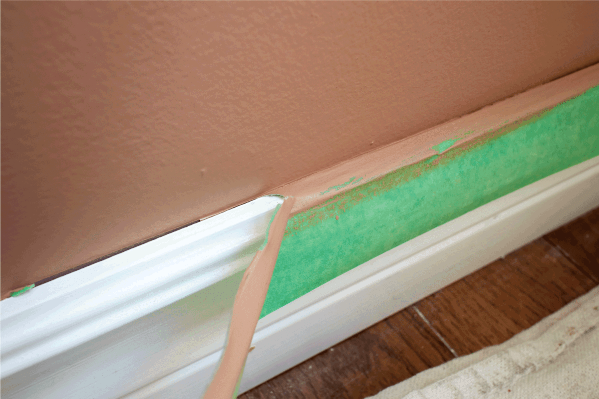 a rip in green painters tape with brown-pink paint on it being removed from white baseboard.