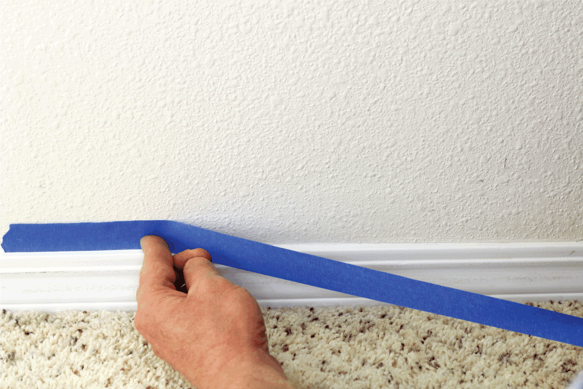 hand preparing to paint wall trim by placing blue painter's tape on the wall above it for protection.